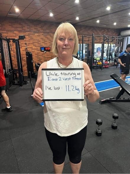 Client holding a whiteboard reading: while training at East 2 West Fitness I've lost 11.2 kg