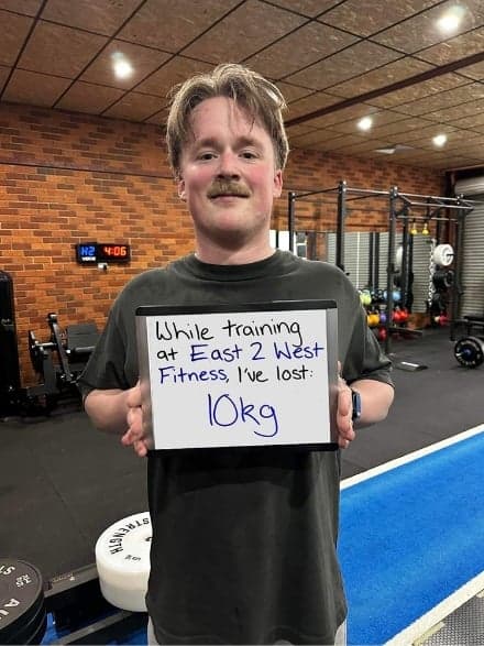 Client holding a whiteboard reading: while training at East 2 West Fitness I've lost 10 kg